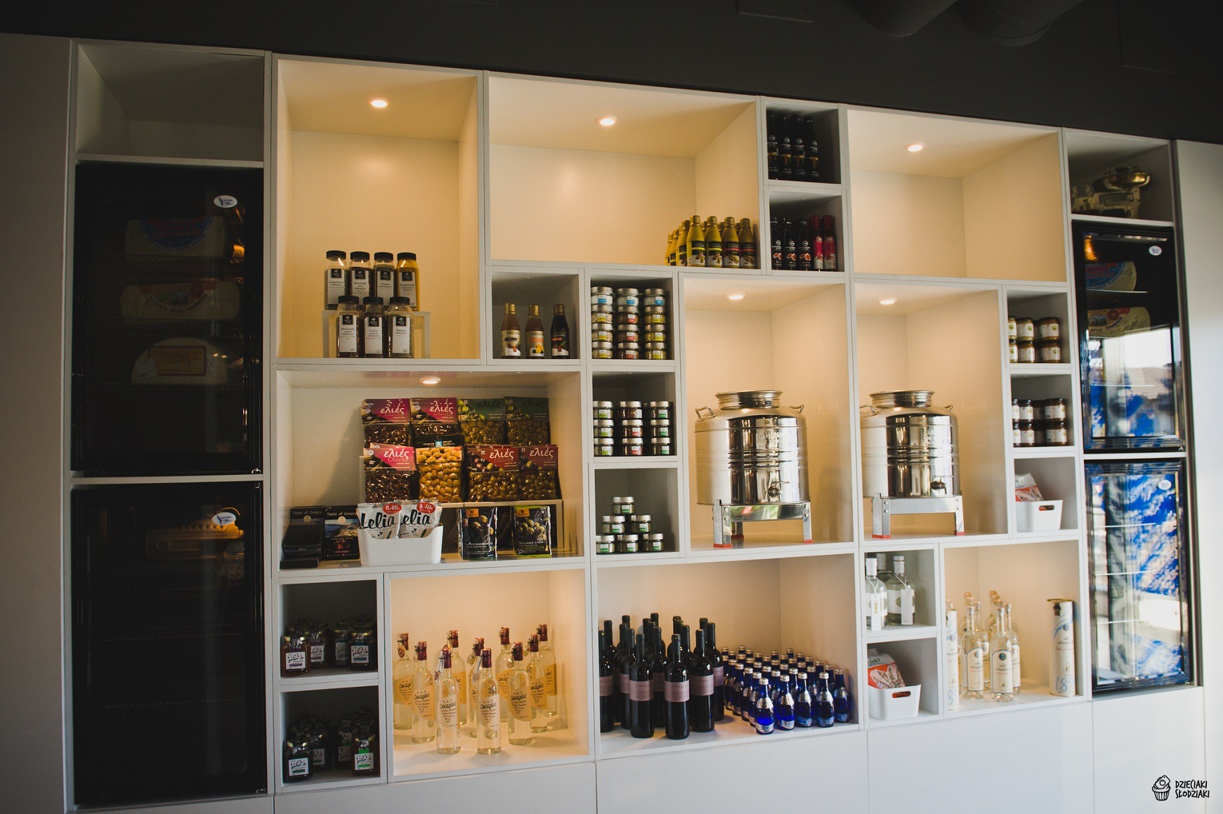 Biała, modułowa półka sklepowa z podświetleniem, prezentująca różnorodne produkty spożywcze: oliwki w opakowaniach, butelki z oliwą, słoiki z przetworami, napoje w szklanych butelkach...