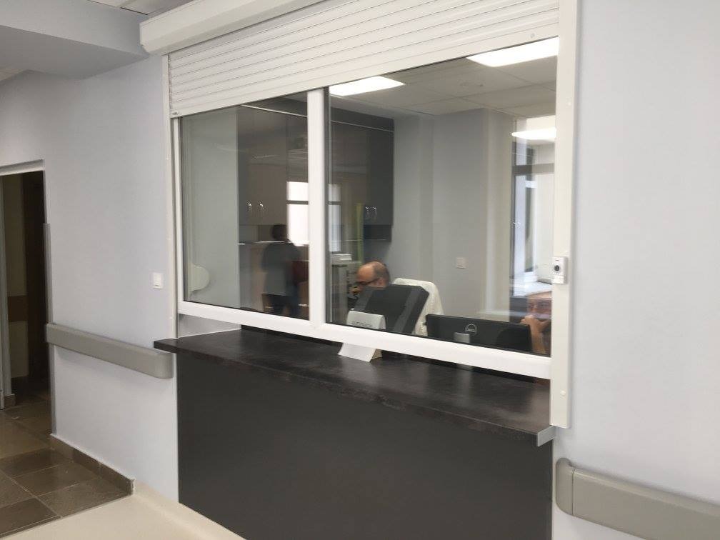 Recepcja z przeszklonym oknem z roletą zewnętrzną, widok na wnętrze biura z pracownikami przy komputerach.