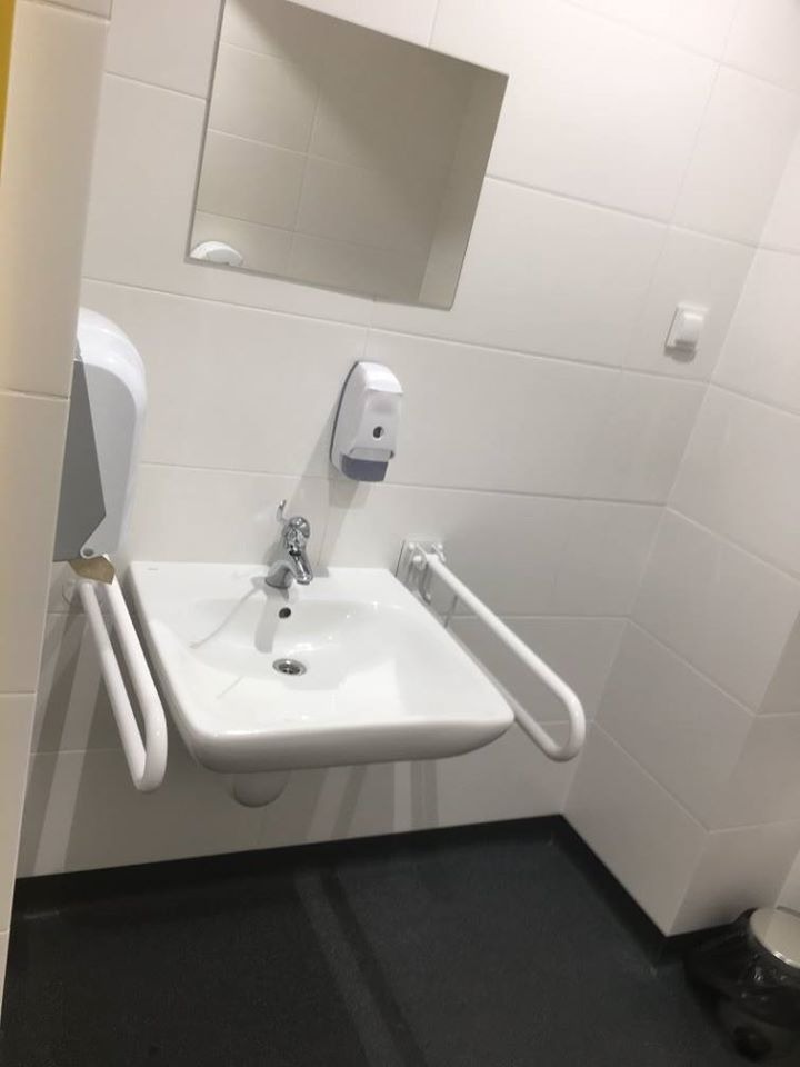 Umywalka dla niepełnosprawnych z poręczami, zamontowana na białej, kafelkowanej ścianie, z lustrem i dozownikiem mydła w tle, na podłodze z ciemnej gumy.