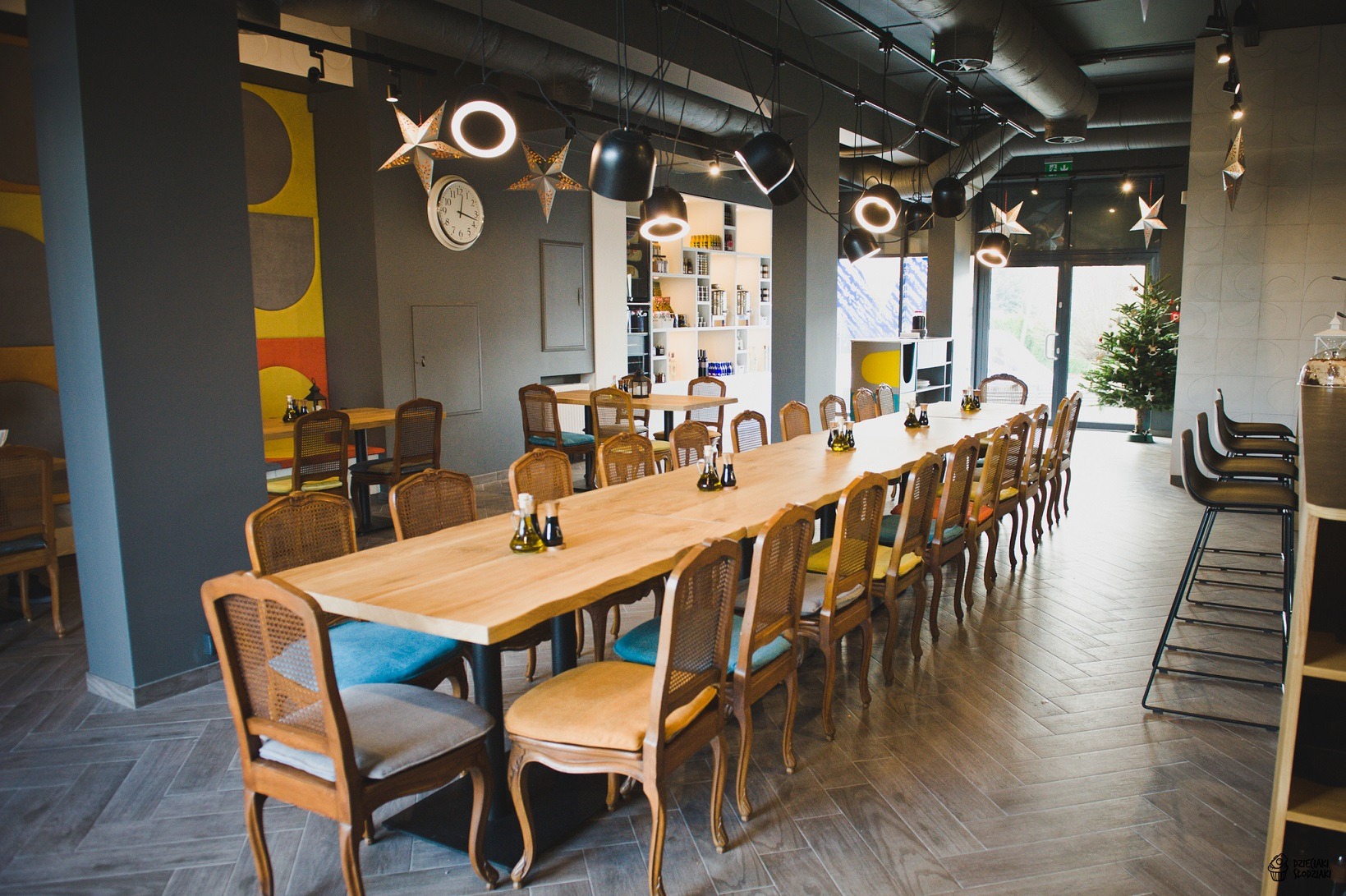 Wnętrze restauracji z długim drewnianym stołem, krzesłami z wiklinowymi oparciami i kolorowymi poduszkami, nowoczesnym oświetleniem oraz dekoracjami w kształcie gwiazd.
