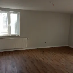 Puste, jasne wnętrze z panelami podłogowymi w odcieniach brązu, białym grzejnikiem pod oknem i szarymi ścianami. Widoczne gniazdko elektryczne przy podłodze i wystające przewody na suficie.