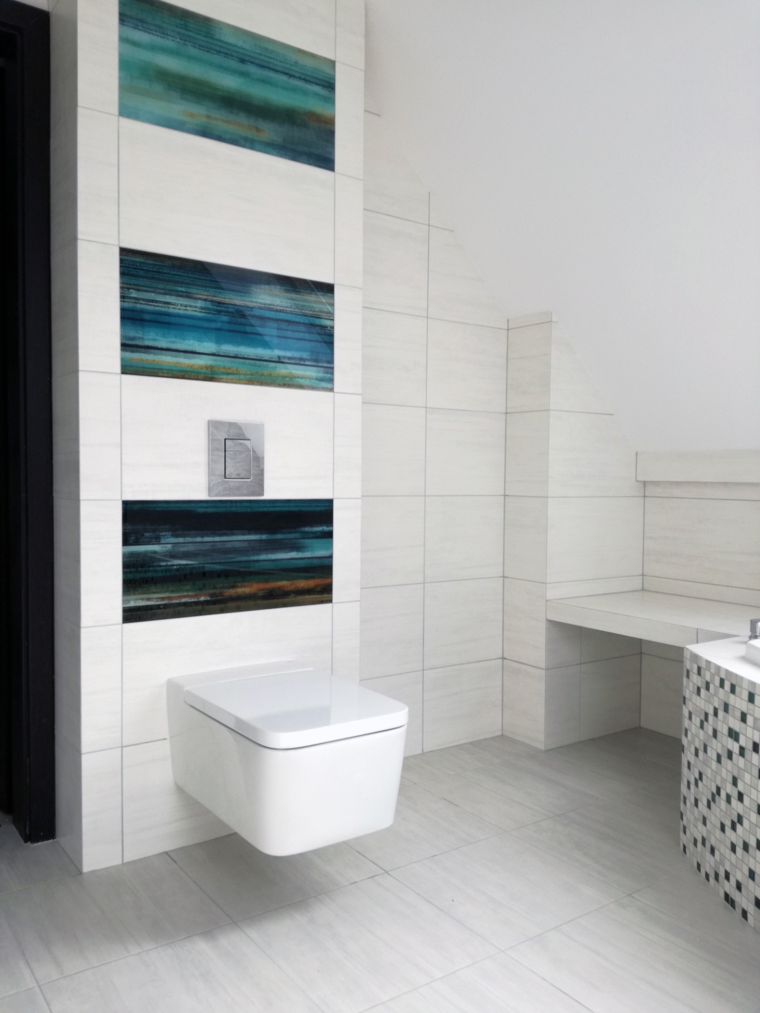 Nowoczesna toaleta podwieszana Geberit w łazience wyłożonej białymi płytkami imitującymi drewno, z dekoracyjnymi panelami w odcieniach turkusu i szarości oraz mozaiką przy umywalce.