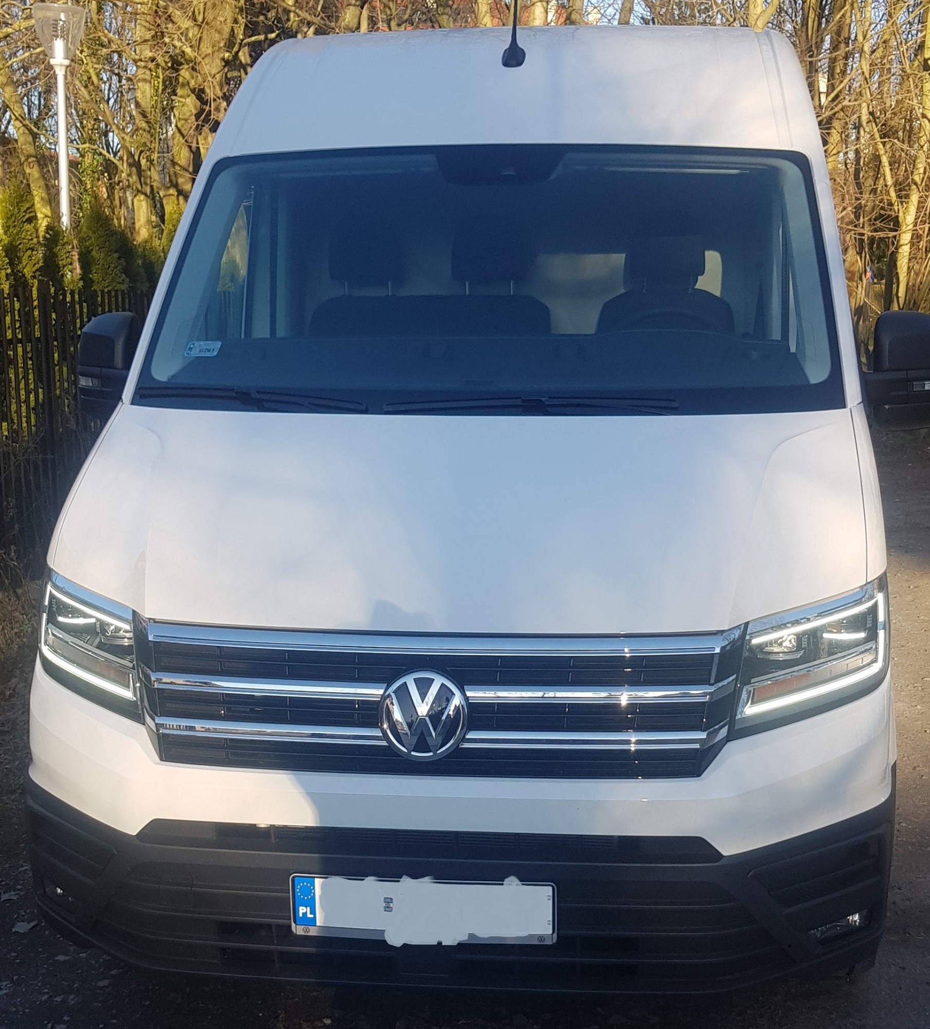 Biały bus marki Volkswagen Crafter, zaparkowany na zewnątrz z widoczną tablicą rejestracyjną z polską flagą na tle drzew bez liści.