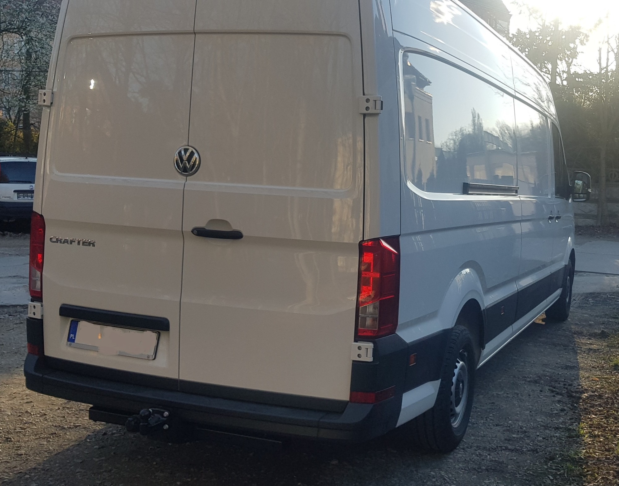 Tył białego samochodu dostawczego Volkswagen Crafter z hakiem holowniczym, zaparkowanego na żwirowej nawierzchni, widoczne światła i tablica rejestracyjna.