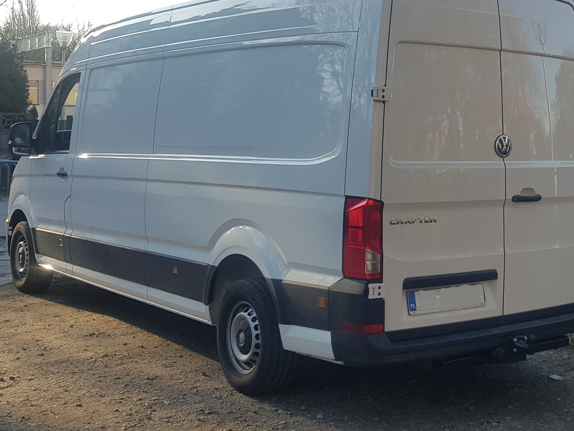 Biały furgon Volkswagen Crafter zaparkowany na żwirowym podłożu, widok z tyłu, z widocznym hakiem holowniczym i tablicą rejestracyjną.