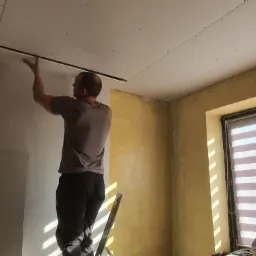 Mężczyzna na drabinie montuje profil sufitowy pod płytę kartonowo-gipsową w pomieszczeniu z ż&oacute;łtą ścianą i oknem z żaluzjami.