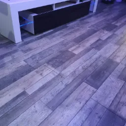 Ułożone panele podłogowe w odcieniach szarości z widocznym fragmentem białej szafki RTV z podświetleniem LED.