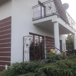 Dwupiętrowy dom z jasną elewacją, brązowymi akcentami i balkonem z ozdobną, metalową balustradą. Na parterze krata z motywem roślinnym, przed budynkiem gęste krzewy.