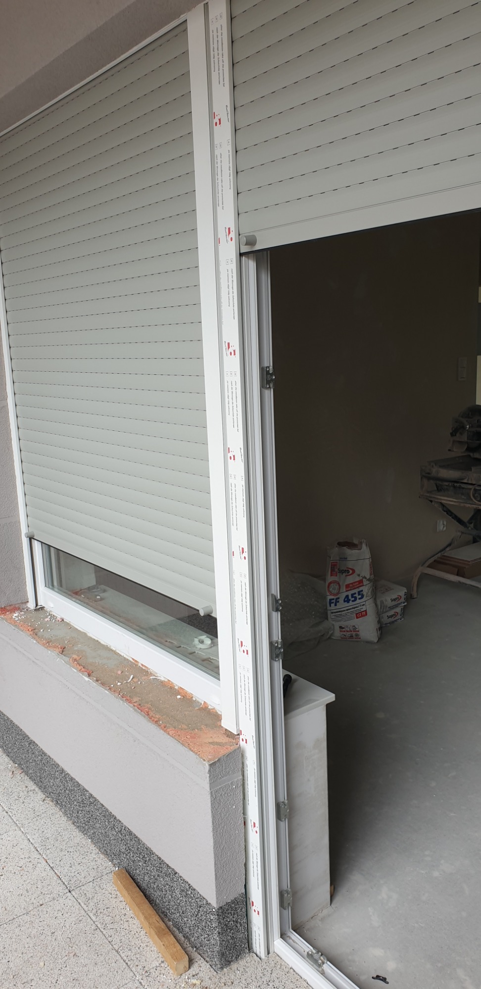Zamknięta roleta zewnętrzna w jasnym odcieniu szarości, zamontowana na oknie z widocznymi elementami montażowymi, w tle worki z zaprawą budowlaną i narzędzia, wskazujące na trwające prace...