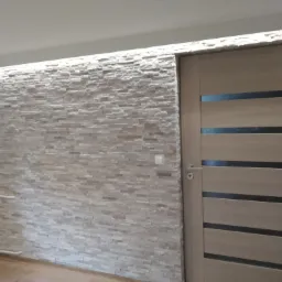 Ściana wewnętrzna wyłożona dekoracyjnym kamieniem, z podświetleniem LED u góry, sąsiadująca z nowoczesnymi drzwiami wewnętrznymi z poziomymi przeszkleniami.