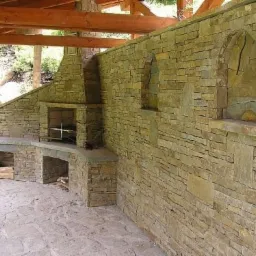 Kamienna zabudowa grilla z kominkiem i blatem roboczym pod zadaszeniem, wykończona nieregularnym kamieniem w odcieniach beżu i szarości, z półką na drewno i kamienną posadzką.