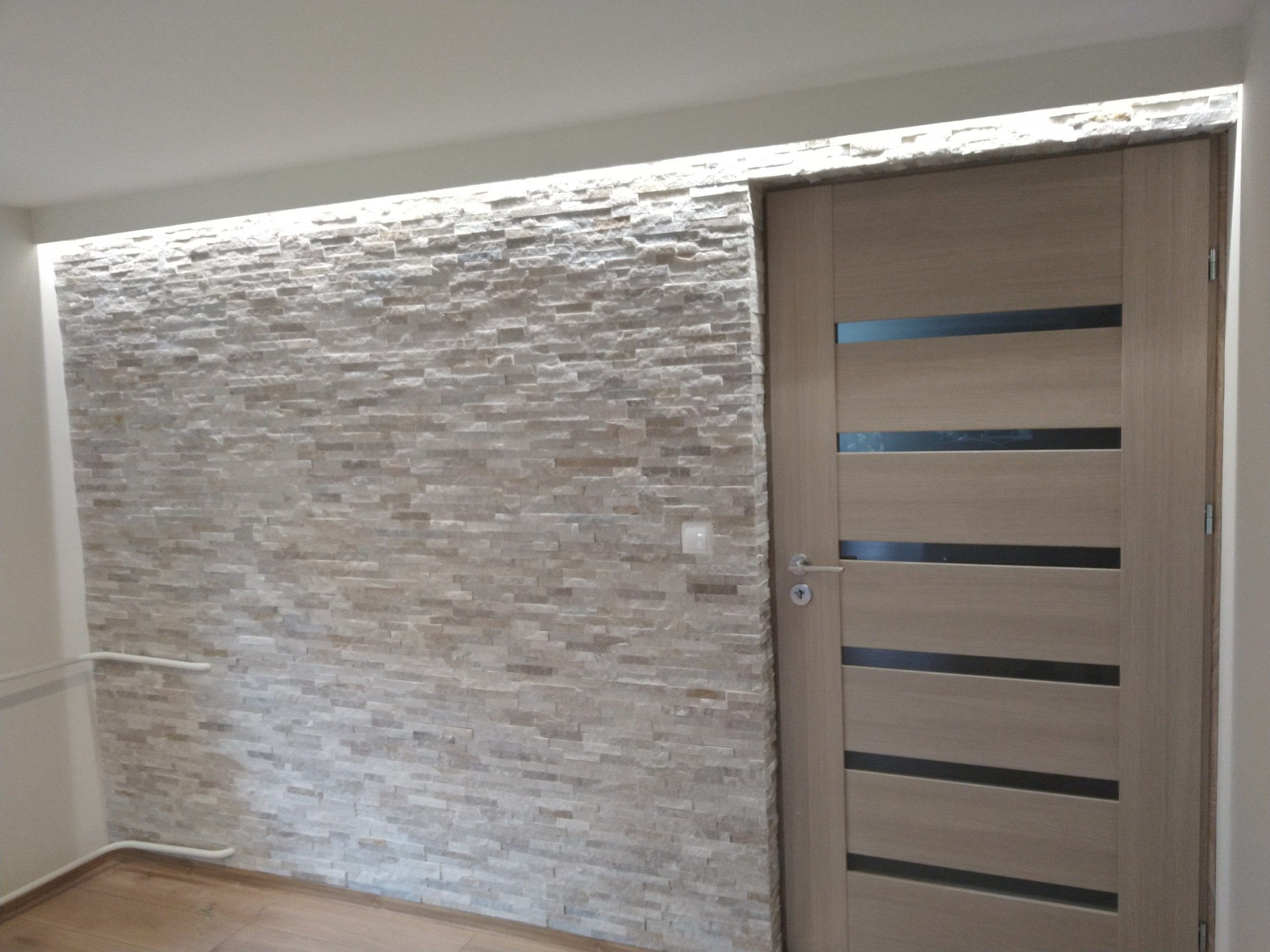 Ściana wewnętrzna wyłożona dekoracyjnym kamieniem, z podświetleniem LED u góry, sąsiadująca z nowoczesnymi drzwiami wewnętrznymi z poziomymi przeszkleniami.