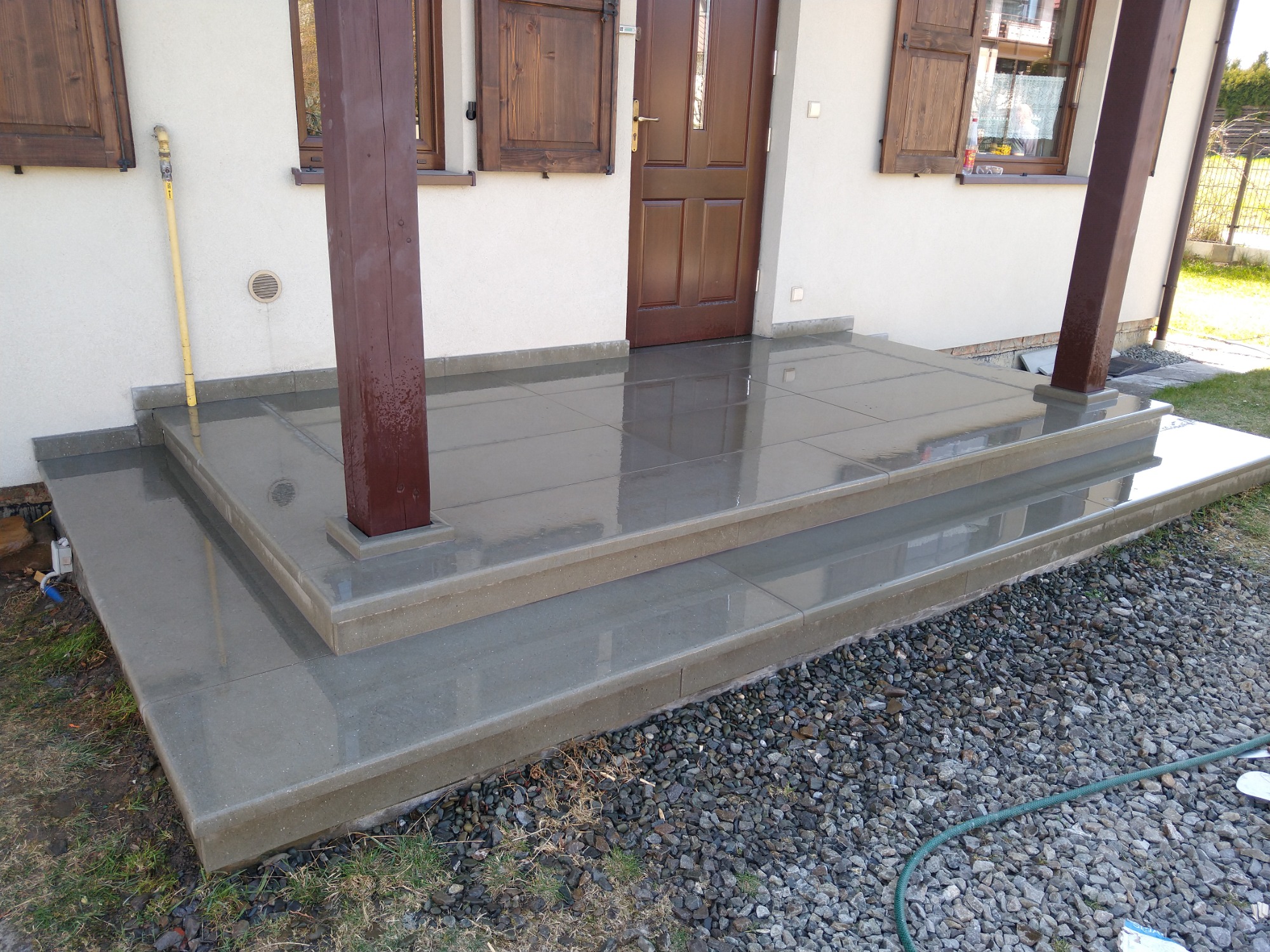 Nowoczesne, dwustopniowe schody wejściowe z szarego kamienia, z brązowymi drewnianymi słupami podtrzymującymi zadaszenie, widok z dołu na drzwi wejściowe i okna z brązowymi okiennicami.