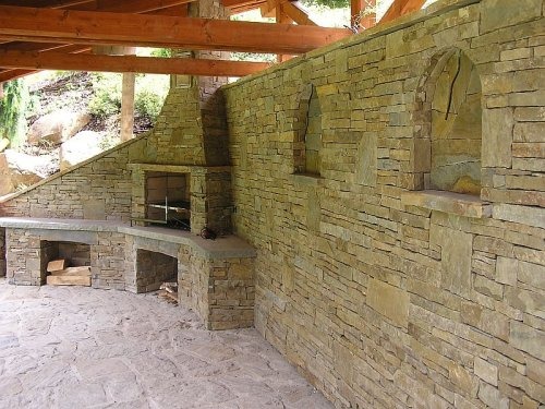 Kamienna zabudowa grilla z kominkiem i blatem roboczym pod zadaszeniem, wykończona nieregularnym kamieniem w odcieniach beżu i szarości, z półką na drewno i kamienną posadzką.