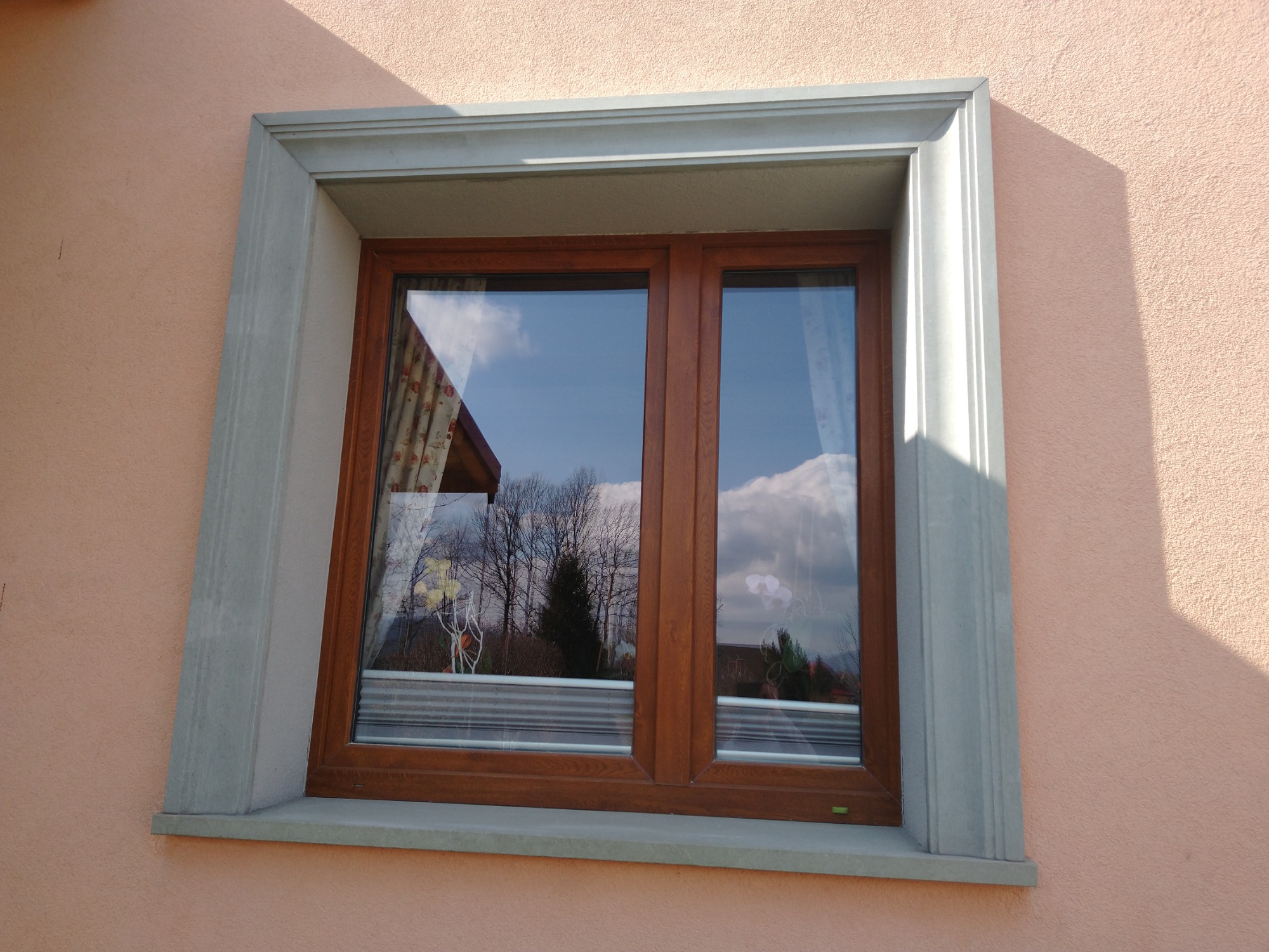Okna z drewnianą ramą i kamiennym obramowaniem na elewacji w kolorze brzoskwiniowym, odbicie nieba i drzew w szybach.