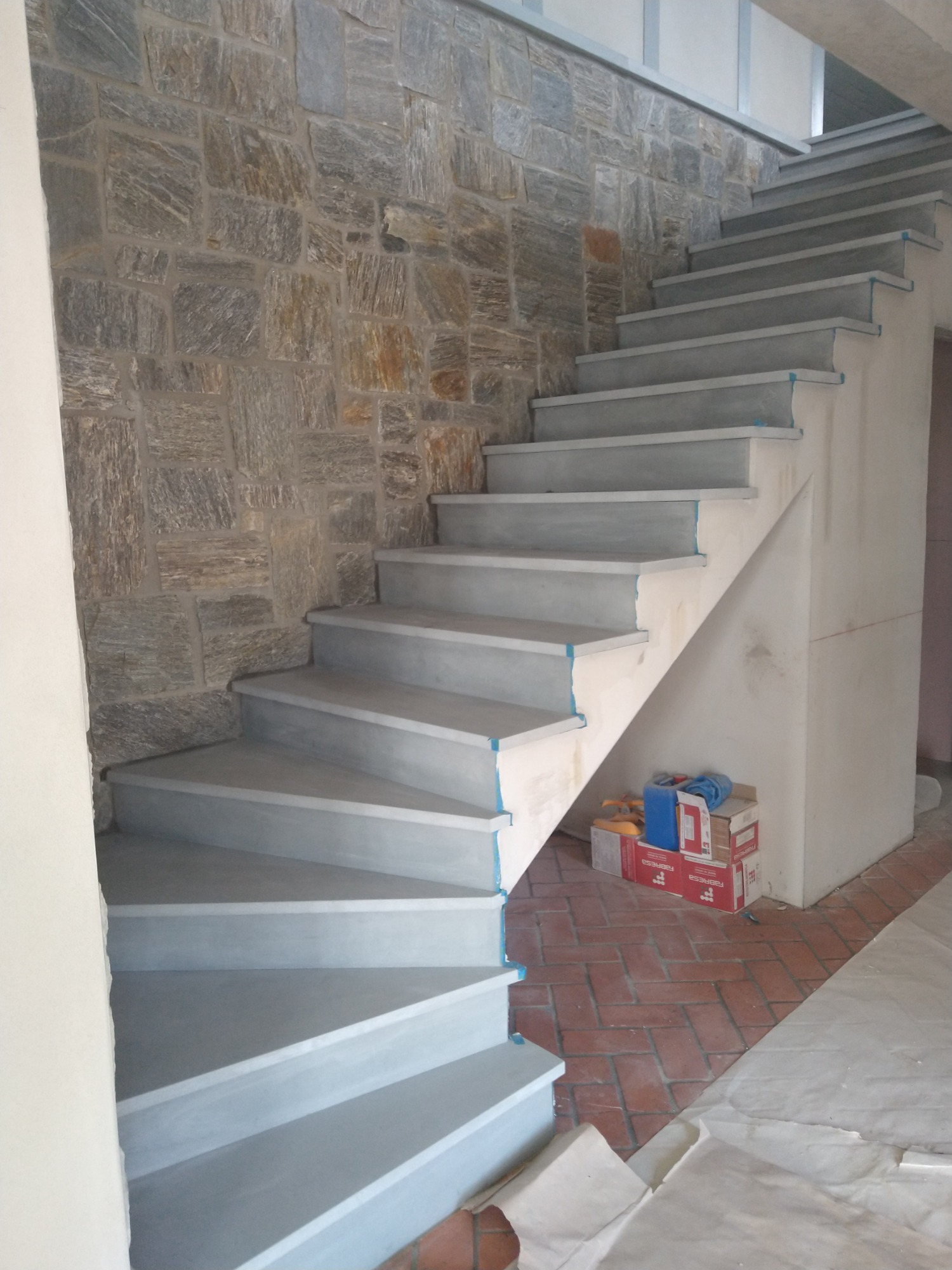 Wewnętrzne schody z szarymi kamiennymi stopniami, ściana wyłożona nieregularnym kamieniem, widoczne zabezpieczenia z taśmy malarskiej na krawędziach stopni, podłoga częściowo przykryta folią...