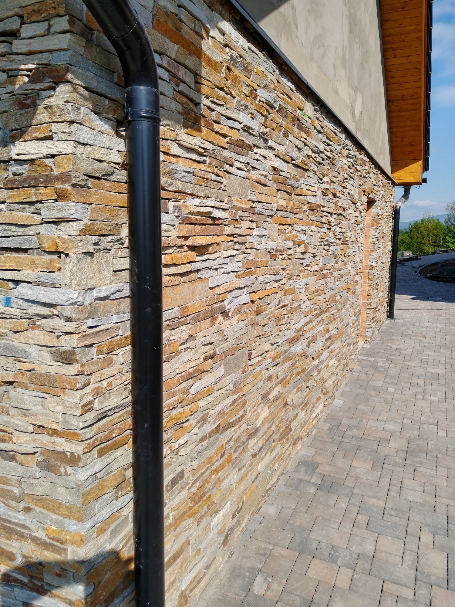 Elewacja budynku wykończona kamieniem naturalnym w odcieniach beżu i szarości, z widoczną czarną rynną spustową i fragmentem chodnika z kostki brukowej.