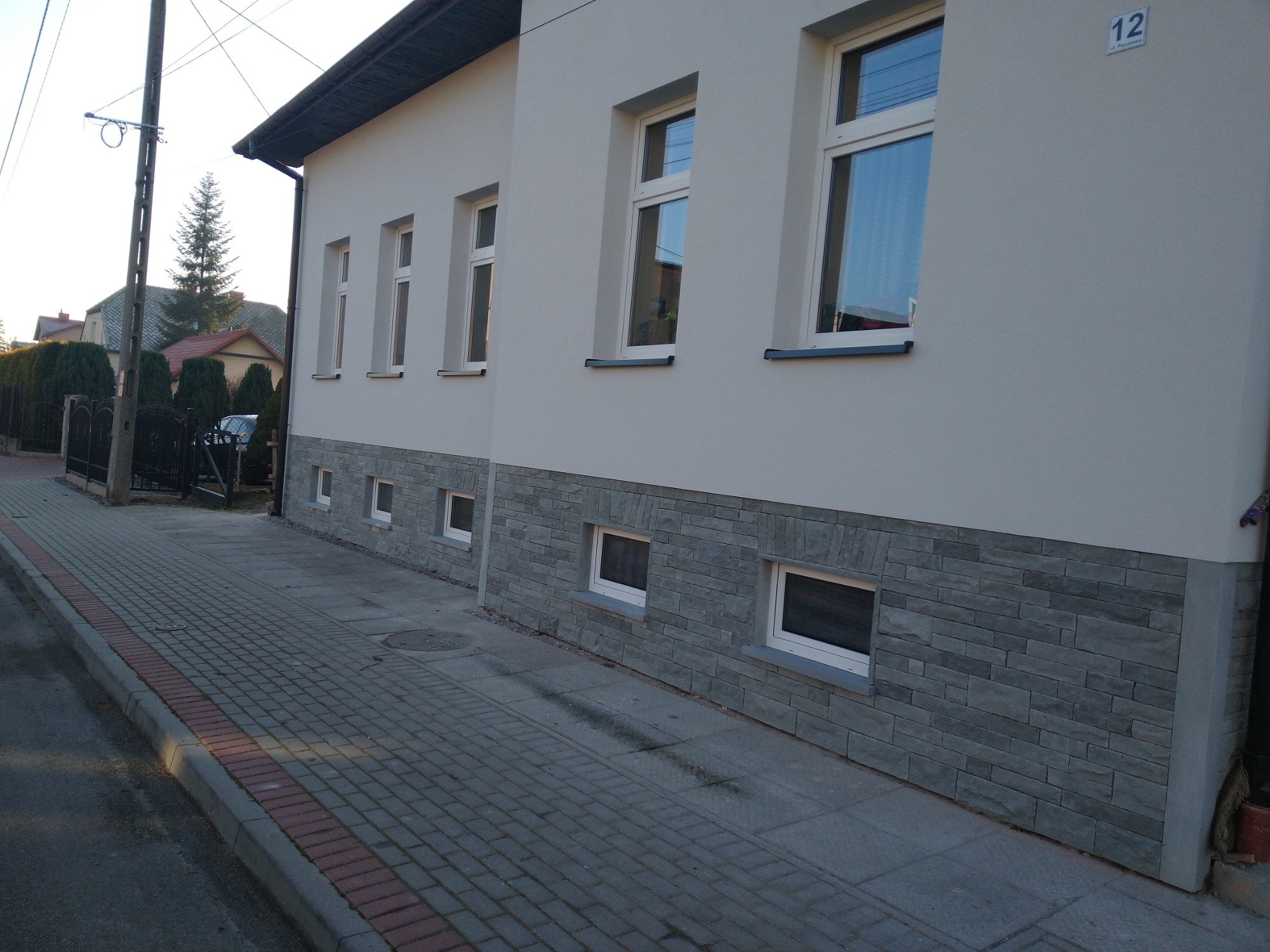 Elewacja budynku z szarą okładziną kamienną na dole, białe okna i jasny tynk. Widoczny chodnik z kostki brukowej i numer domu '12'.