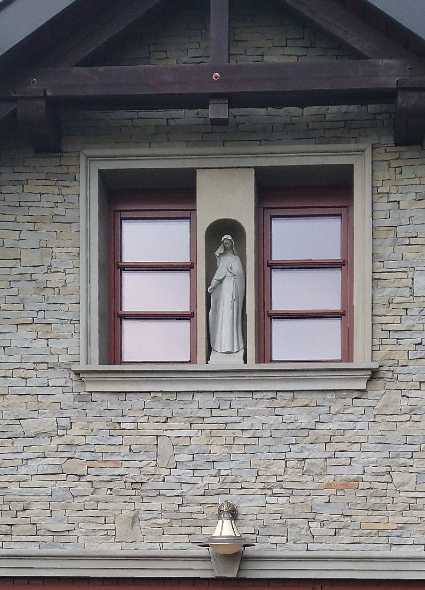 Fragment elewacji budynku z oknem flankowanym wnęką z figurą Matki Boskiej, wykonanej z kamienia w odcieniach szarości, z widoczną lampą elewacyjną poniżej.