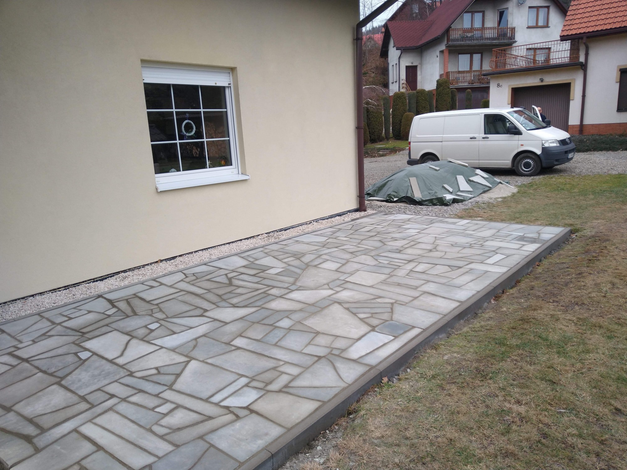 Ułożony taras z nieregularnych płyt kamiennych, obok budynku z beżową elewacją i białym oknem, w tle domy i zaparkowany biały van.