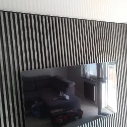 Nowoczesna ściana z pionowych paneli dekoracyjnych w kolorze szaro-czarnym, odbicie wnętrza w lustrzanej powierzchni z widoczną sofą, skrzynkami narzędziowymi i oknem.