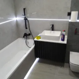 Nowoczesna łazienka w szarościach z białą wanną, umywalką nablatową na czarnej szafce z ryflowanym frontem i czarną armaturą, oświetlona listwą LED wzdłuż ściany, widoczne środki czystości i papier...