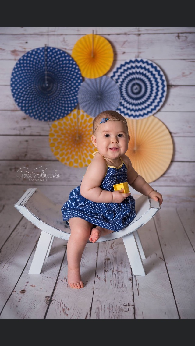 Uśmiechnięta niemowlęca dziewczynka w granatowej sukience w białe kropki, siedząca na białej ławce z żółtym klockiem w ręku, tło z drewnianych desek i dekoracyjnych, kolorowych rozet.