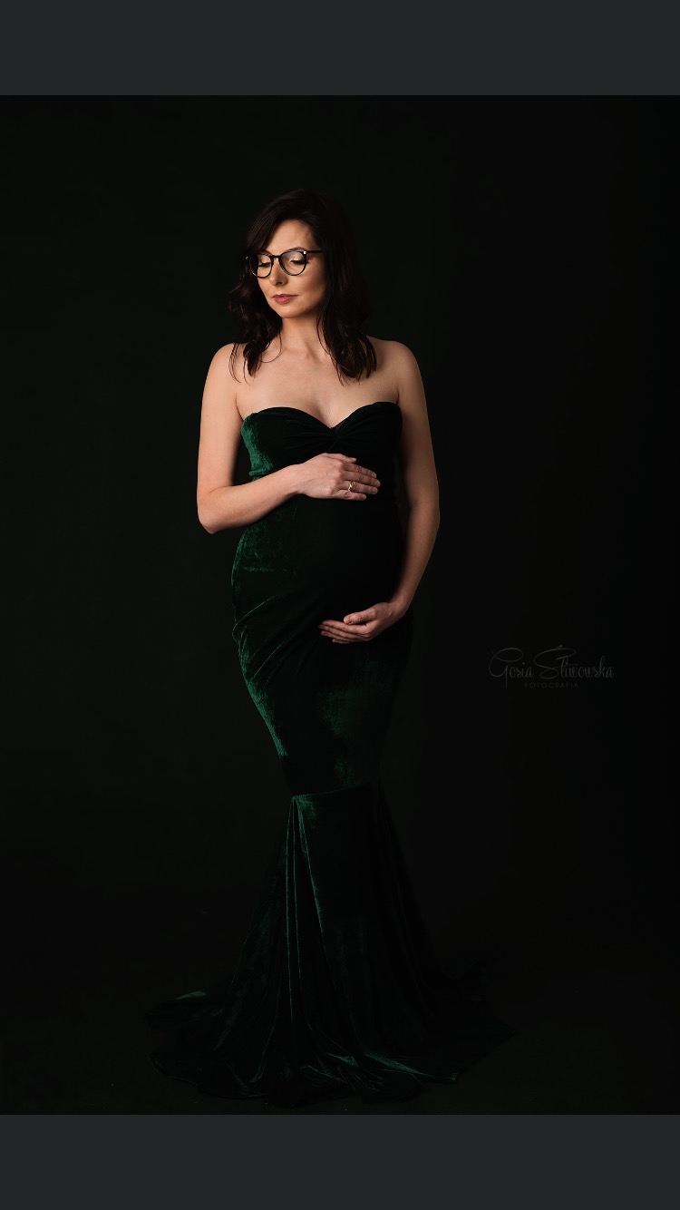 Kobieta w ciąży w szmaragdowej, aksamitnej sukni bez ramiączek, z rękami na brzuchu i okularami na twarzy, pozuje na ciemnym tle.