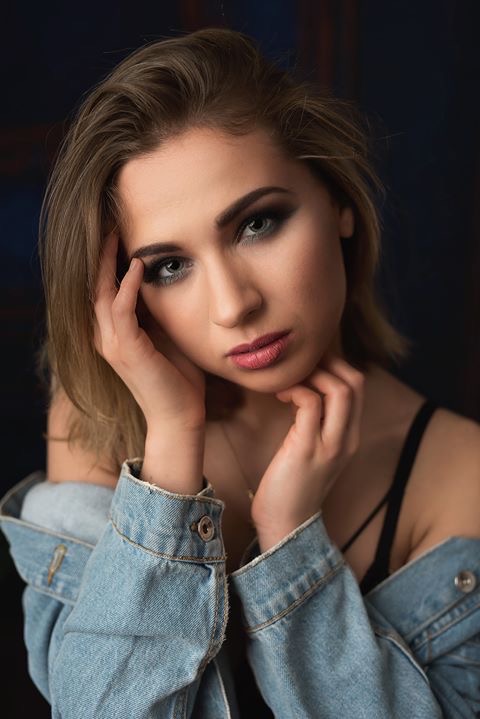 Portret kobiety z mocnym makijażem oczu, w dżinsowej kurtce, z rękami przy twarzy, na ciemnym tle.