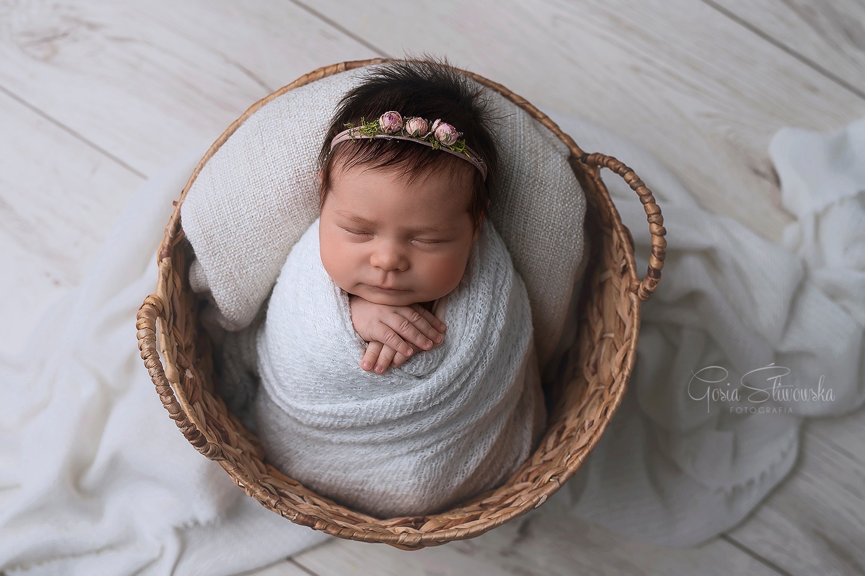 Noworodek zawinięty w biały kocyk, śpiący w wiklinowym koszu z małą opaską z różowymi kwiatami na głowie, widok z góry.