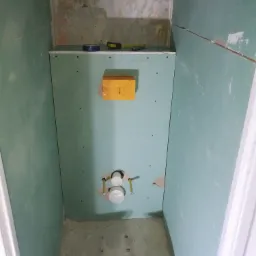 Wnętrze małej łazienki w trakcie prac wykończeniowych; widoczne płyty gipsowo-kartonowe na ścianach, przygotowane otwory na instalację sanitarną oraz robocze obuwie na podłodze.