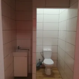 Wykończona łazienka z białymi płytkami ściennymi, drewnopodobną podłogą, toaletą i małą umywalką z szafką w minimalistycznym stylu.