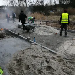 Prace przygotowawcze do ułożenia nawierzchni w Skrzynkach: robotnicy rozładowują piasek z taczek obok ułożonych obrzeży, widoczny sprzęt budowlany i narzędzia.