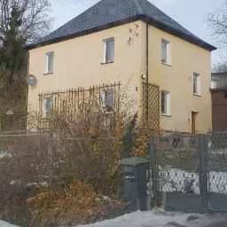 Dwupiętrowy dom z ciemnym, skośnym dachem z blachy trapezowej, widoczny zza ogrodzenia i krzewów w zimowej scenerii.