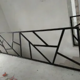Metalowa balustrada schodowa z geometrycznym wzorem, czarna rama, surowe wykończenie wnętrza, widoczny fragment niezakończonej ściany.