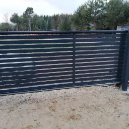 Automatyczna brama przesuwna z poziomych profili, widoczny mechanizm zębatkowy, w tle las i częściowo samochód.