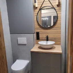 Nowoczesna łazienka z drewnianymi panelami na ścianie, okrągłym lustrem na sznurku, umywalką nablatową w odcieniach szarości i szafką pod umywalką w kolorze grafitowym, wisząca toaleta.