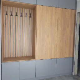 Szara zabudowa meblowa z drewnianymi akcentami: szafa z panelami imitującymi drewno i otwartą wnęką z wieszakami, widoczna na tle szarej podłogi w trakcie remontu.