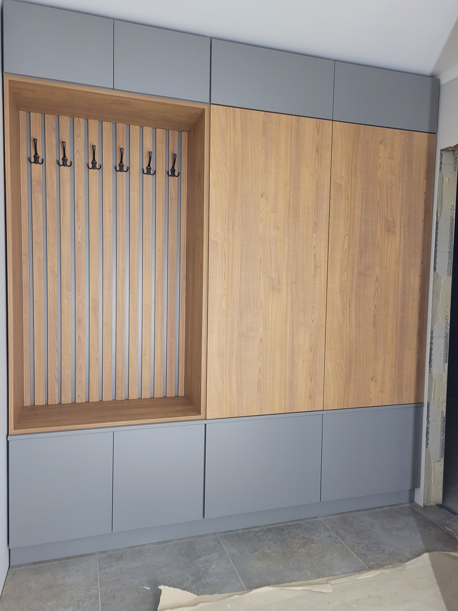 Szara zabudowa meblowa z drewnianymi akcentami: szafa z panelami imitującymi drewno i otwartą wnęką z wieszakami, widoczna na tle szarej podłogi w trakcie remontu.