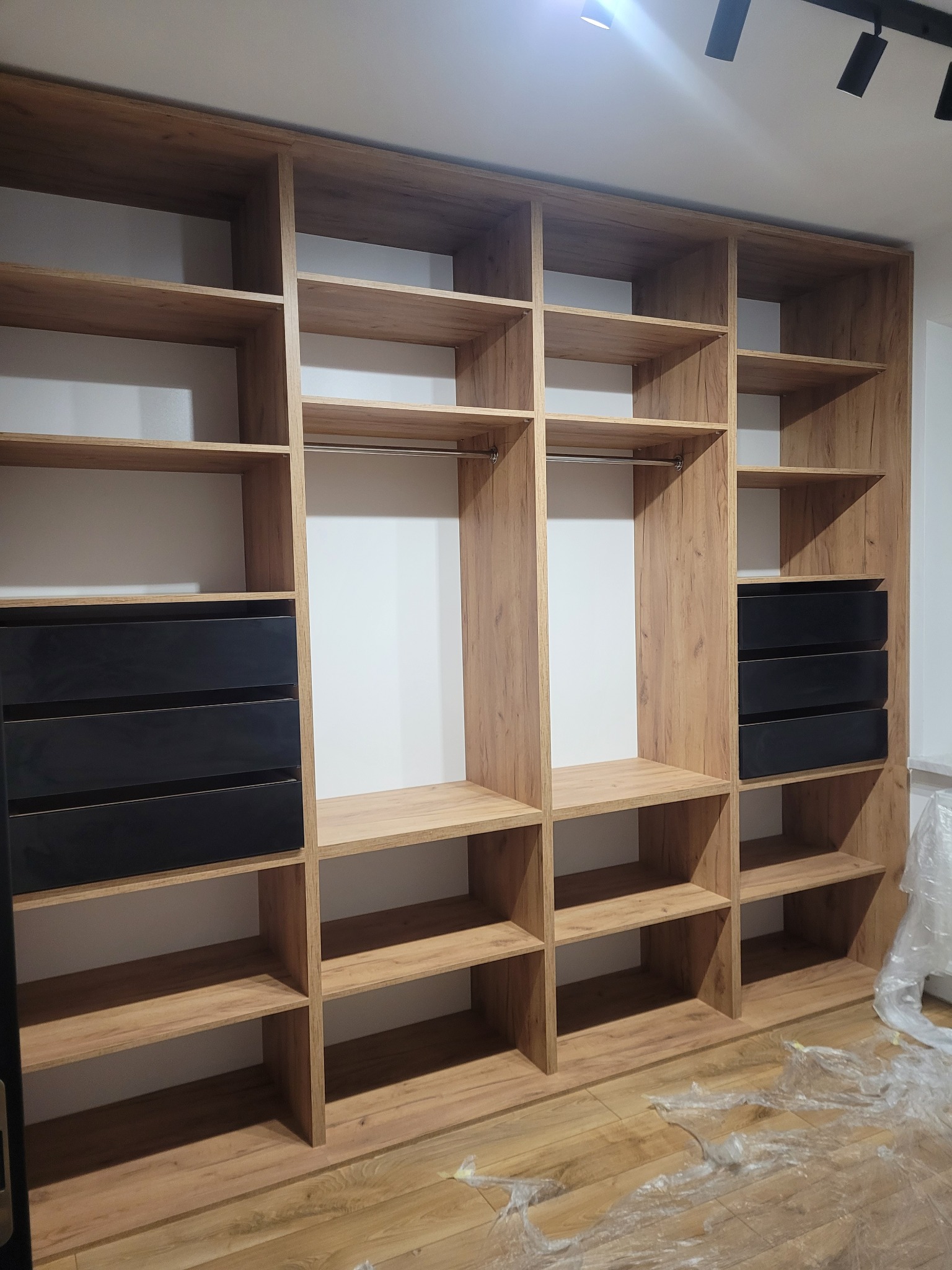 Wnętrze szafy wnękowej z drewnianymi półkami i czarnymi szufladami, widoczne elementy montażowe i folia ochronna na podłodze.