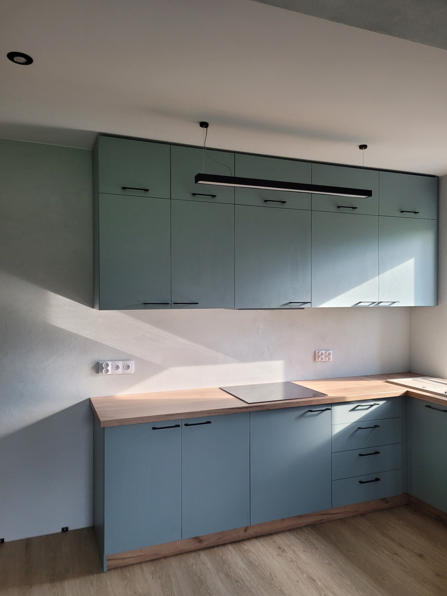 Nowoczesna, błękitna zabudowa kuchenna z drewnianym blatem, oświetlona podłużną lampą LED, z widocznymi gniazdkami elektrycznymi na ścianie i fragmentem drewnianej podłogi.