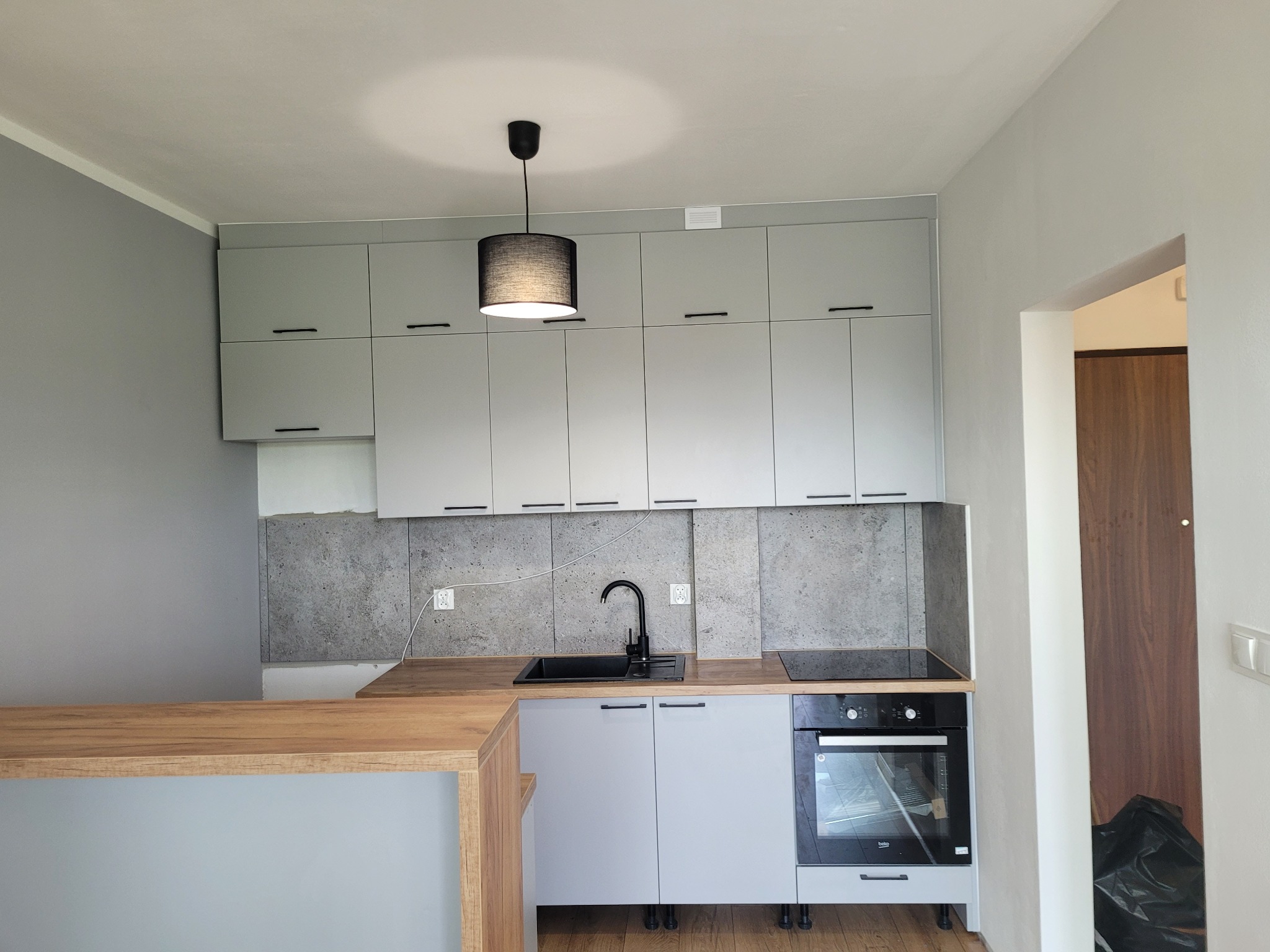 Jasnoszara kuchnia z drewnianym blatem i czarną armaturą, oświetlona lampą z abażurem, z szarymi płytkami imitującymi kamień na ścianie.