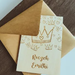 Drewniana kartka z wygrawerowanym napisem 'Roczek Emilki' i motywem koron, leżąca na brązowej kopercie i ciemnym drewnianym tle.