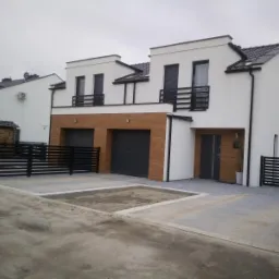 Nowoczesne domy murowane z drewnianą okładziną garaży, czarnymi balustradami i ogrodzeniem, widoczne niedokończone prace brukarskie na podjeździe.