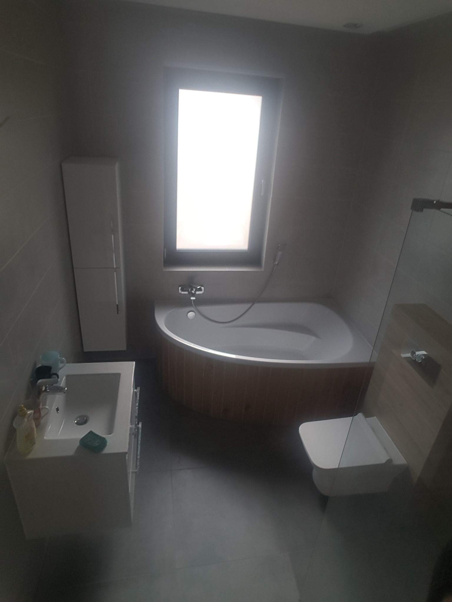 Nowoczesna łazienka w szarościach z narożną wanną obudowaną drewnem, wiszącą szafką umywalkową i toaletą.