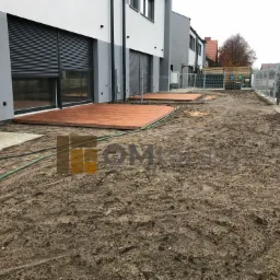 Dwa tarasy z desek kompozytowych przed nowoczesnym budynkiem w trakcie prac wykończeniowych ogrodu, widoczne ogrodzenie i przygotowany teren pod trawnik.