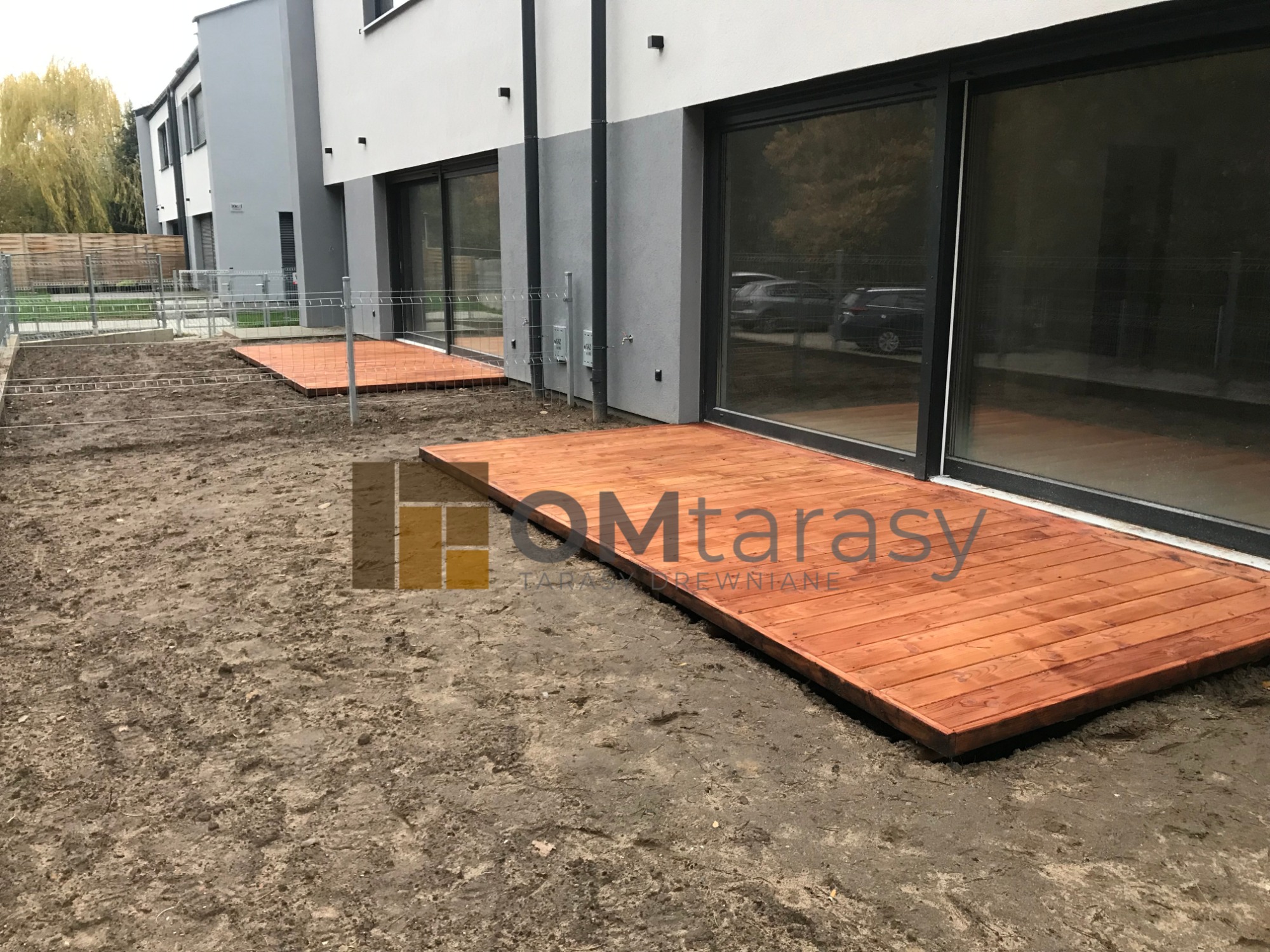 Dwa tarasy z desek w kolorze ciepłego brązu, przylegające do nowoczesnego domu z dużymi przesuwnymi oknami, teren wokół w trakcie przygotowania, widoczne ślady ziemi.