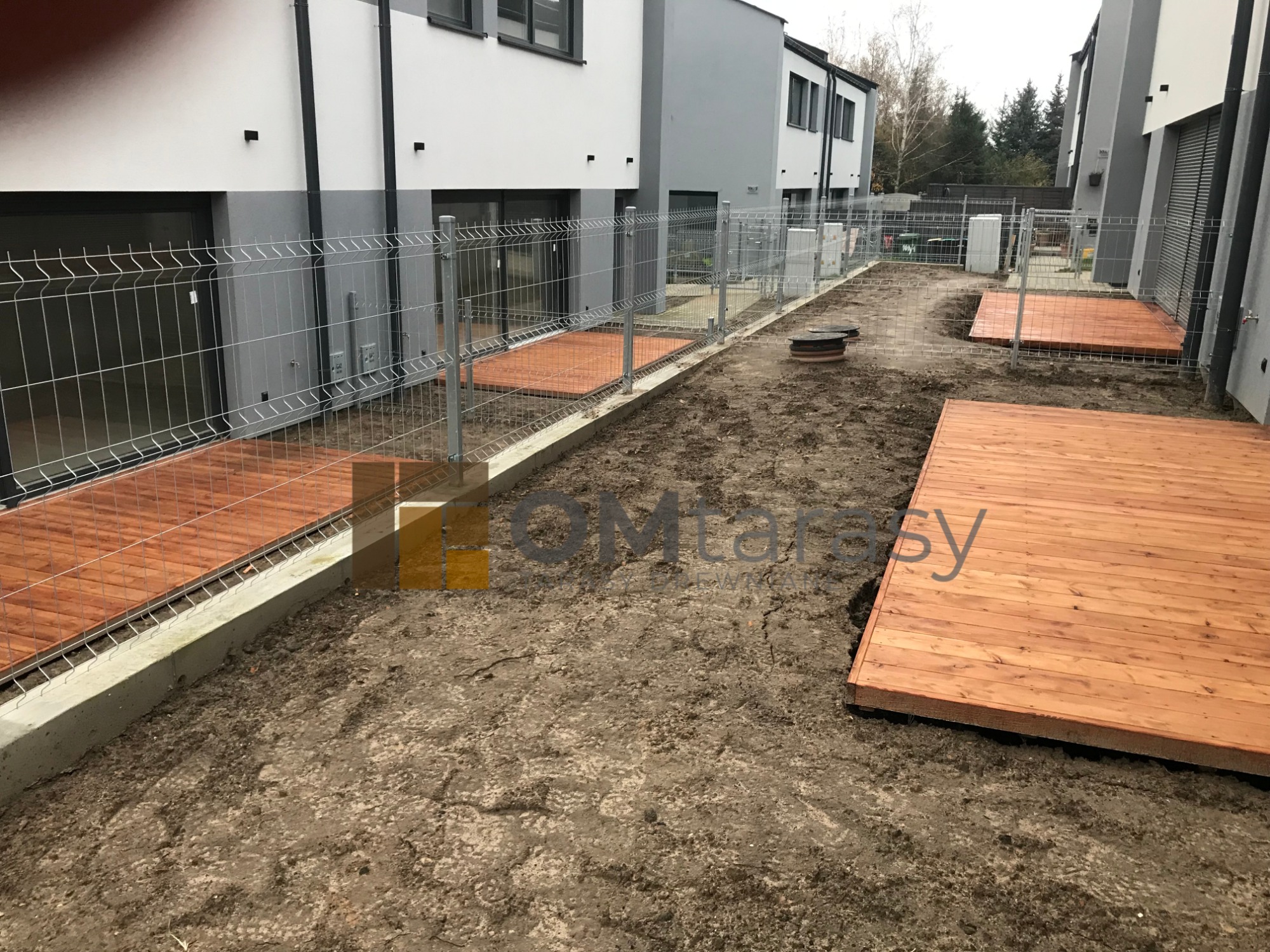 Montaż kilku tarasów z desek w ogrodzie szeregowca, widoczne ogrodzenie z siatki i przygotowany teren.