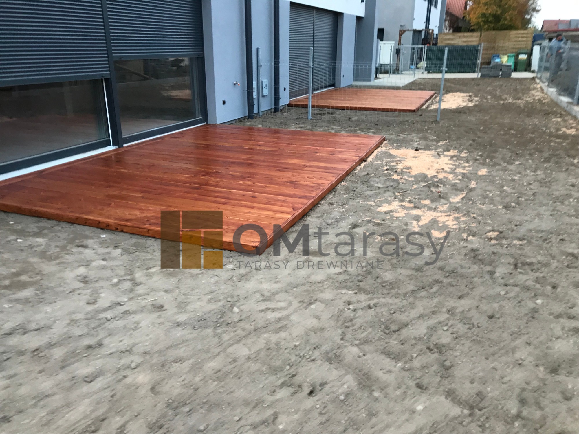 Dwa prostokątne, świeżo zaimpregnowane tarasy z desek, przylegające do nowoczesnego domu z szarymi roletami zewnętrznymi, teren wokół w trakcie prac wykończeniowych.