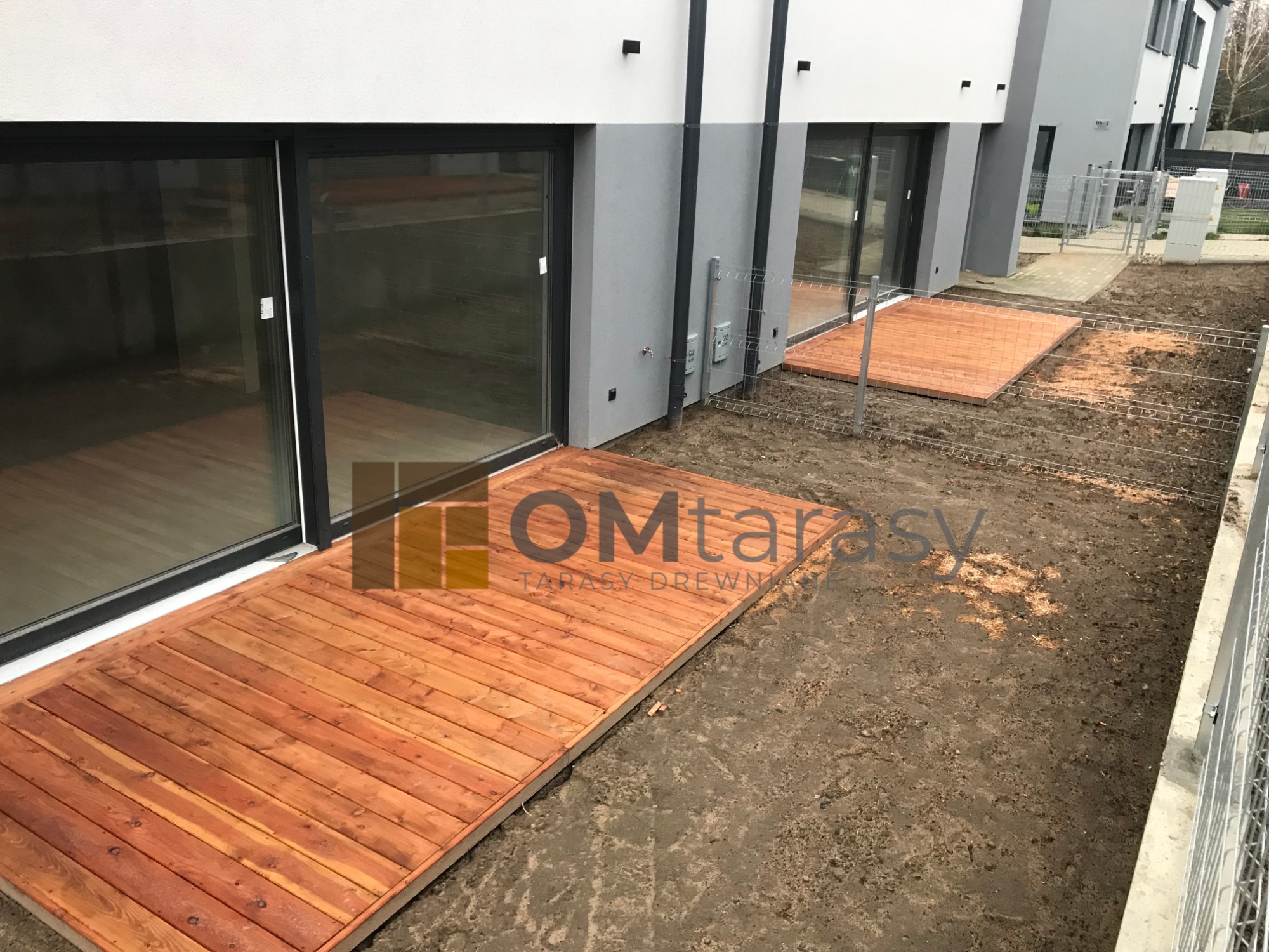 Dwa nowe, prostokątne tarasy z desek z jasnego drewna, przylegające do nowoczesnego, szarego budynku z dużymi przesuwnymi oknami, teren wokół tarasów jeszcze niezagospodarowany, widoczna metalowa...